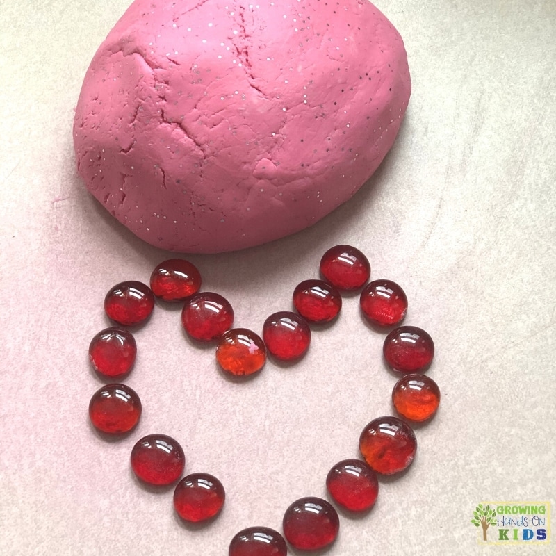 red valentine themed play dough with red glass beads shaped into a heart on a white counter background. 