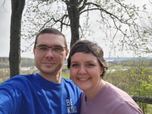 Picture of a man in a blue sweatshirt with brown hair and glasses next to a women in a pink shirt with short brown hair taking a selfie at an overlook in the woods by the Ohio river.