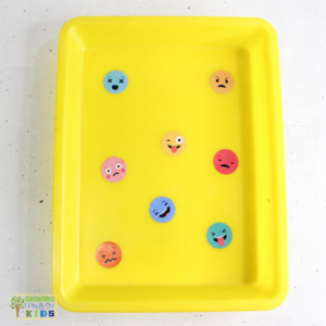 This image shows a yellow tray with various colorful emoji face stickers placed inside. Each emoji displays a different emotion, such as happy, sad, surprised, angry, and playful. The tray is part of a sensory activity designed to help children recognize and explore different emotions through visual aids. The simple, clean setup focuses on emotional expression and is likely intended for a fun, interactive learning activity.