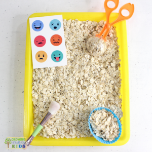 This image shows a sensory play activity setup with a yellow tray filled with dry oats. A pair of orange scoop scissors and a small brush are included, along with a blue cupcake liner filled with oats. On the left side, there is a card featuring various colorful emoji faces expressing different emotions, such as happy, sad, angry, and surprised. This setup is intended for a tactile sensory experience, helping children explore textures and emotions through play.