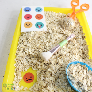 This image shows a sensory play activity setup with a yellow tray filled with dry oats. A pair of orange scoop scissors and a small brush are included, along with a blue cupcake liner filled with oats. On the left side, there is a card featuring various colorful emoji faces expressing different emotions, such as happy, sad, angry, and surprised. This setup is intended for a tactile sensory experience, helping children explore textures and emotions through play.