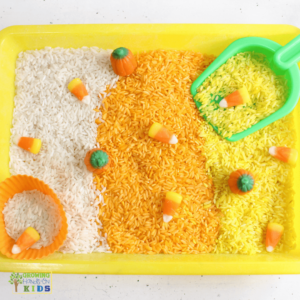 Yellow sensory bin filled with layers of white, orange, and yellow rice, decorated with candy corn and candy pumpkins, alongside a green scoop and an orange silicone cup for scooping.