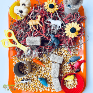 Farm animal sensory bin setup featuring plastic farm animals, corn kernels, sunflowers, shredded paper, and small toys on an orange tray for hands-on sensory play.
