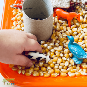 Child's hand playing with a miniature cow in a farm animal sensory bin filled with corn kernels, toy animals, and a cardboard tube on an orange tray.