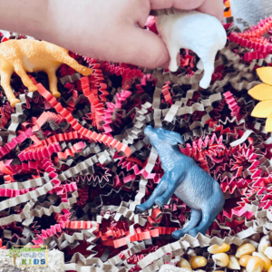 Child's hand playing with a miniature sheep in a farm animal sensory bin filled with corn kernels, toy animals, and fall paper shreds.