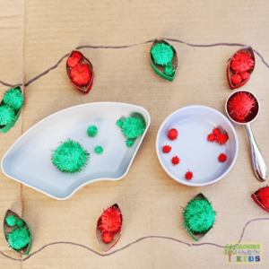 Christmas-themed activity setup featuring red and green glitter pom poms in white bowls, a metal scoop with red pom poms, and painted toilet paper roll strips resembling Christmas lights glued onto cardboard with a drawn string connecting them.