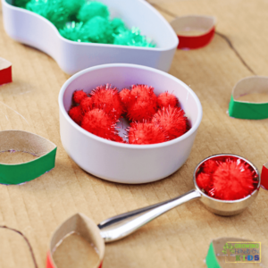 Christmas-themed activity setup featuring red and green glitter pom poms in white bowls, a metal scoop with red pom poms, and painted toilet paper roll strips resembling Christmas lights glued onto cardboard with a drawn string connecting them.