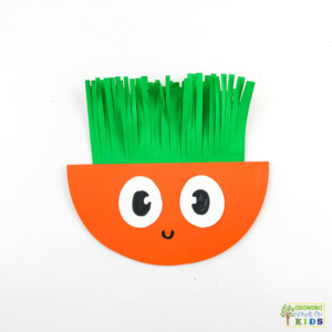 A colorful paper craft of a rocking carrot made from orange and green construction paper, featuring a happy face with large, cartoonish eyes. The carrot's leafy top is made of fringed green paper, and the background is white.