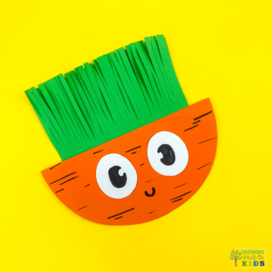 A colorful paper craft of a rocking carrot made from orange and green construction paper, featuring a happy face with large, cartoonish eyes. The carrot's leafy top is made of fringed green paper, and the background is bright yellow.