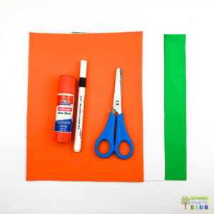 Supplies needed for making a rocking carrot craft. Supplies include orange, white, and green construction paper, glue stick, black marker, blue scissors, all on a white background.