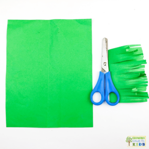 A colorful paper craft of a rocking carrot made from orange and green construction paper. The photo shows green paper in a square, blue scissors, and a green rectangle of paper with fringe.