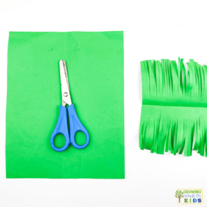 A colorful paper craft of a rocking carrot made from orange and green construction paper. The photo shows green paper in a square, blue scissors, and a green rectangle of paper with fringe.