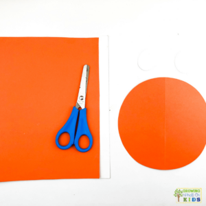 A colorful paper craft of a rocking carrot made from orange and green construction paper. The photo shows orange paper in a square, a orange circle, and blue scissors.