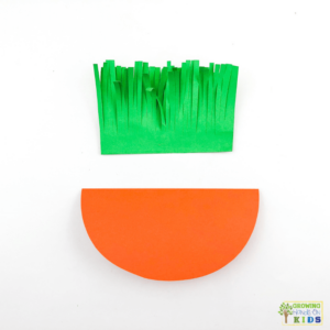 A colorful paper craft of a rocking carrot made from orange and green construction paper. The carrot's leafy top is made of fringed green paper.