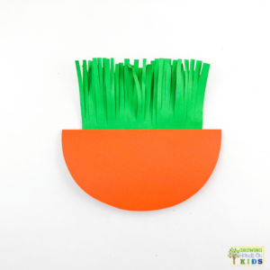 A colorful paper craft of a rocking carrot made from orange and green construction paper. The carrot's leafy top is made of fringed green paper.