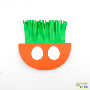 A colorful paper craft of a rocking carrot made from orange and green construction paper, featuring white eyes. The carrot's leafy top is made of fringed green paper, and the background is white.