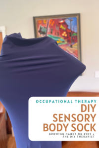 A child is inside a navy blue sensory body sock, stretching the fabric outward with their arms. The background shows a warmly lit room with a colorful painting of a street scene on the wall. A white and orange text overlay in the bottom right corner reads: “Occupational Therapy DIY Sensory Body Sock – Growing Hands-On Kids + The DIY Therapist.