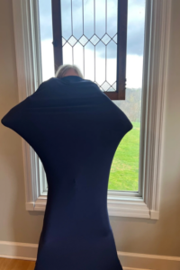 A child is inside a dark blue sensory body sock, stretching the fabric outward with their arms and legs, creating a star-like shape. They are standing in front of a large window with a view of green grass and a cloudy sky.