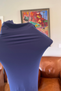 A child is inside a navy blue sensory body sock, stretching the fabric outward with their arms, forming a diamond shape. The background features a tan wall with a colorful framed painting of a street scene and a brown leather chair.