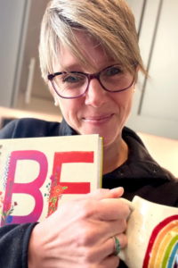 A picture of Kerry from The DIY Therapist, with short blond hair and purple glasses holds a colorful book with the large word "BE" on the cover and a cream-colored mug with a rainbow handle. She is wearing a black top and standing in a warmly lit kitchen with gray cabinets in the background.
