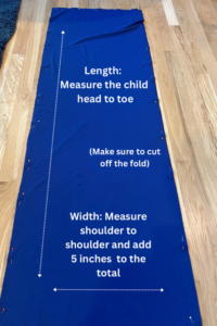 A piece of dark blue fabric laid out on a wooden floor with pins along the edges. White text on the fabric provides measuring instructions for making a body sock. It says: "Length: Measure the child head to toe (Make sure to cut off the fold)." At the bottom it says: "Width: Measure shoulder to shoulder and add 5 inches to the total." Dotted arrows indicate direction for measuring length (top to bottom) and width (left to right).