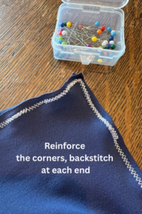 A close-up of a sewn corner of dark blue stretch fabric on a wooden table. The seam is finished with a zigzag stitch. A clear plastic box filled with colorful sewing pins is in the background. White text on the fabric reads: "Reinforce the corners, backstitch at each end.