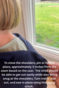 A child with a short blond bob haircut is wearing a partially sewn navy blue sensory body sock near a window with a green yard outside. The fabric is draped over their shoulders with a visible pin holding it in place. White instructional text on the image reads: “To close the shoulders, pin or hold in place, approximately 3 inches from the seam based on the user. The child should be able to get out easily while also being snug at the shoulders. Turn inside out, and sew in place using the zigzag stitch.