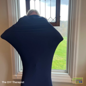 A child is inside a dark blue sensory body sock, stretching the fabric outward with their arms and legs, creating a star-like shape. They are standing in front of a large window with a view of green grass and a cloudy sky. The text "The DIY Therapist" appears in the bottom left corner, and a "Growing Hands-On Kids" logo is in the bottom right.