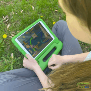 Child sitting on grass using a tablet in a green case, playing an interactive map-based reading game in the Reading Eggs app. The screen shows colorful characters and a path with numbered levels.