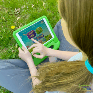Child sitting on the grass using a tablet in a green case to navigate the Reading Eggs app. The screen displays different program options including Reading Eggspress, Mathseeds, and the Library section.