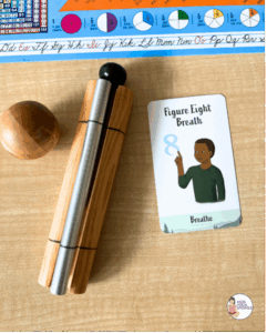 A classroom desk with a chime instrument, wooden mallet, and a "Figure Eight Breath" card featuring an illustrated child tracing a sideways 8 with their finger. A colorful educational chart and alphabet strip are visible in the background. Card is from Kids Yoga Stories.