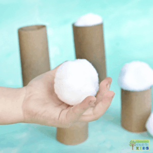 Child’s hand holding a white pom pom snowball in front of cardboard tubes of different heights, demonstrating a winter fine motor balance activity.