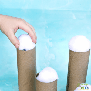 Child placing a white pom pom snowball on top of a cardboard tube, practicing balance and fine motor control in a winter-themed activity.
