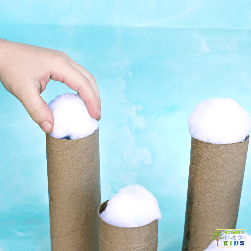 Child placing a white pom pom snowball on top of a cardboard tube, practicing balance and fine motor control in a winter-themed activity.