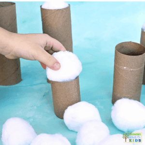 Child balancing a white pom pom snowball on a cardboard tube while other pom poms rest nearby, working on fine motor and hand-eye coordination skills.