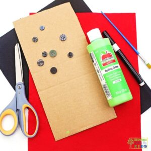 Supplies for a ladybug button fine motor activity including cardboard, green paint, black and red felt, buttons, scissors, marker, and paintbrush laid out on a surface.